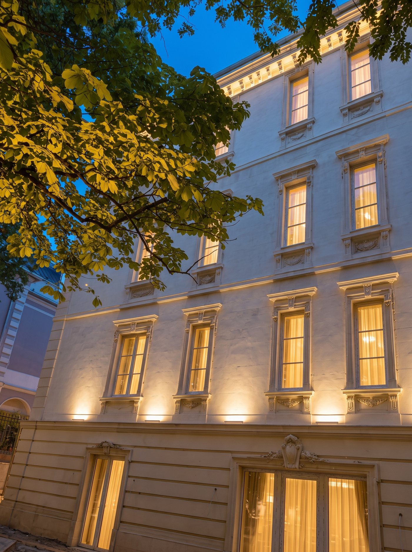 The cream neoclassical façade of Stefánia út 99 at dusk, every window lit, under a chestnut tree